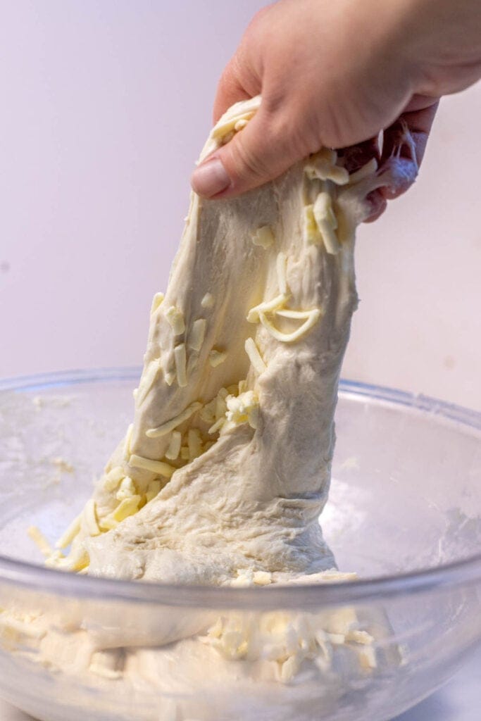 A hand reaching into a bowl of dough and stretching the dough upwards with grated butter in the dough.