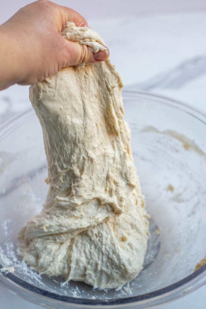 A hand reaching into the bowl and grabbing the dough and stretching it upwards. 
