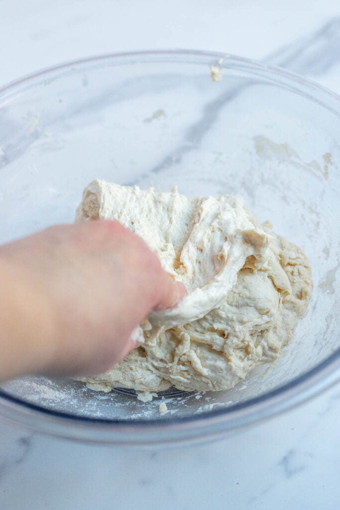 A hand pulling the stretch dough back on itself in a glass bowl.