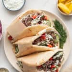 three beef gyros on a plate surrounded by condiments to top the gyros.