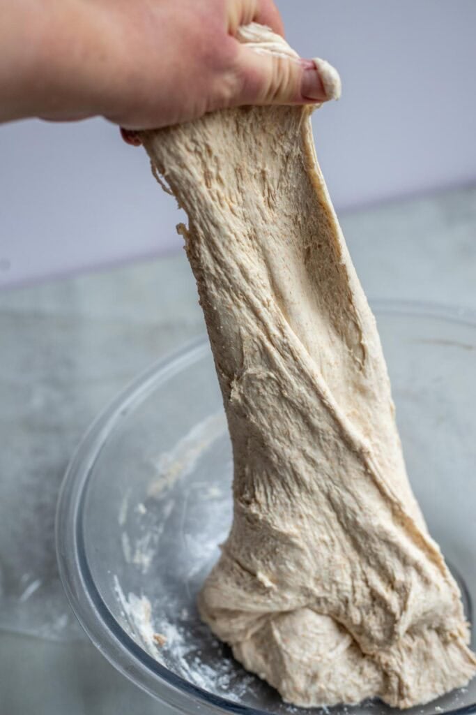A hand pulling dough up out of a glass bowl for a stretch and fold.