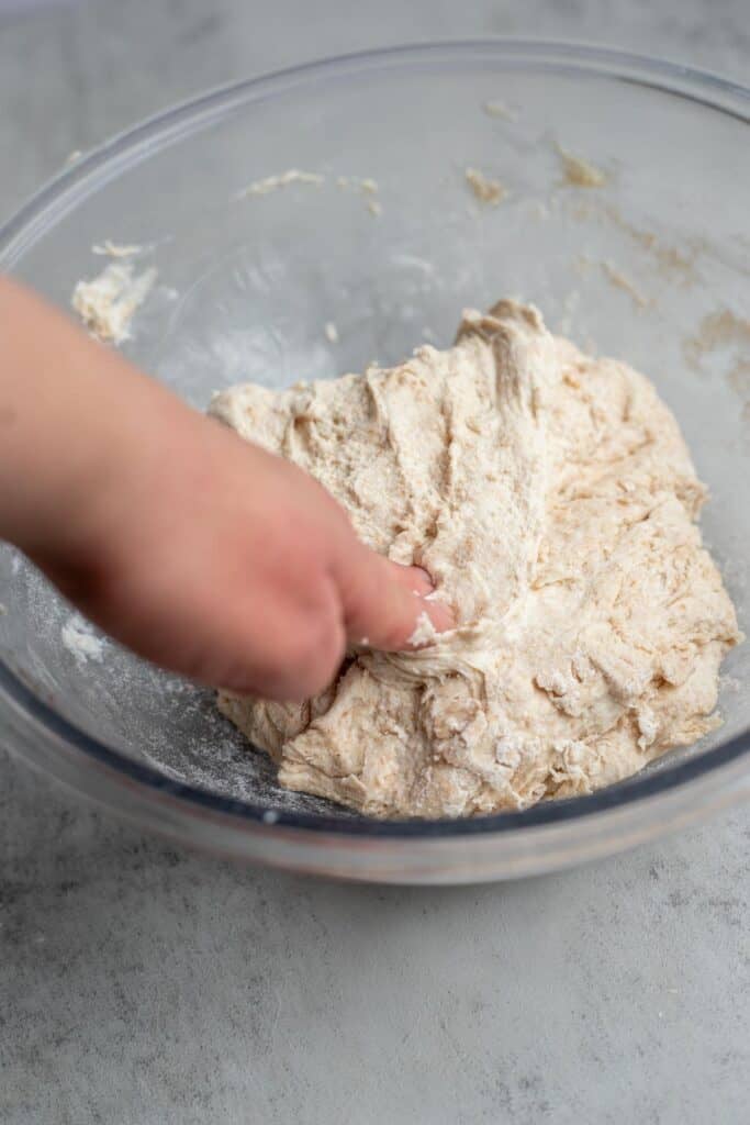 A hand pressing dough down into a glass bowl as part of the stretch and fold process.