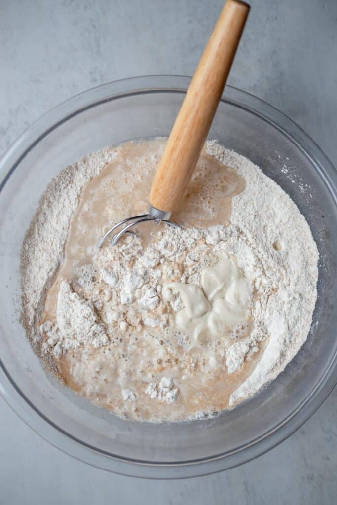 A glass bowl with flour and sourdough starter and a dough whisk.