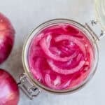 a glass pint jar full of red fermented onions with two whole onions beside the jar.