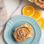 one orange cinnamon roll on a blue plate, surrounded by decorative orange slices.