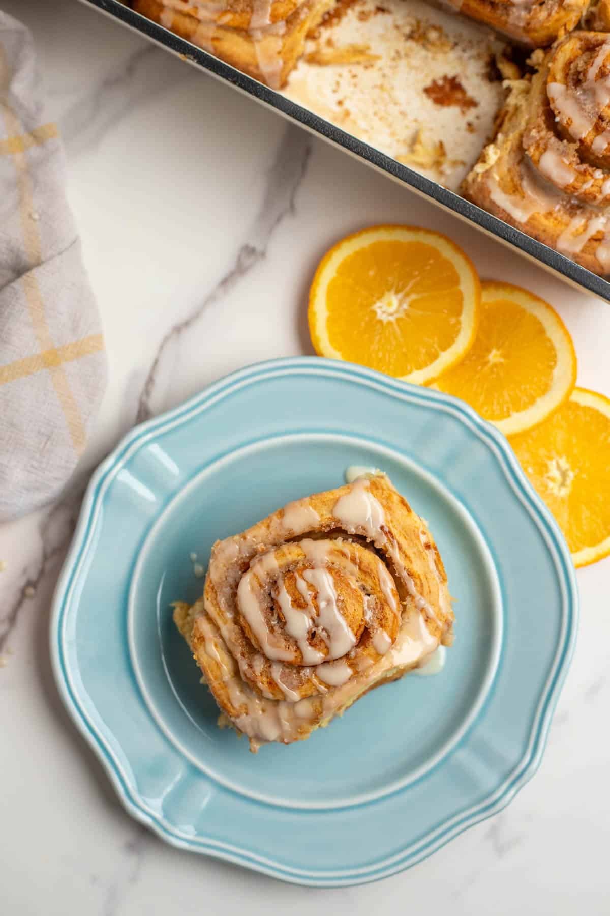 One orange cinnamon roll on a blue plate, surrounded by decorative orange slices. 