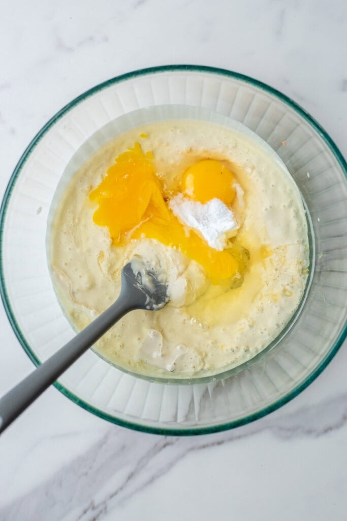 Sourdough starter, eggs, salt, and baking soda in a glass mixing bowl with a spoon to stir.
