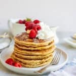 a stack of whole wheat pancakes topped with butter, whipped cream, raspberries, and powdered sugar.