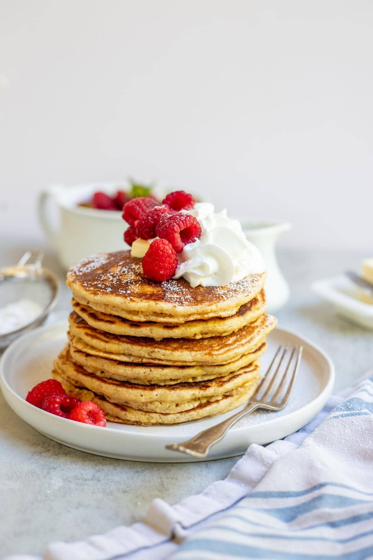 A stack of whole wheat pancakes topped with butter, whipped cream, raspberries, and powdered sugar.