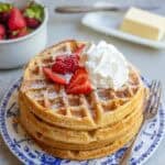 a stack of whole wheat waffles on a blue decorative plate topped with whipped cream and sliced strawberries.