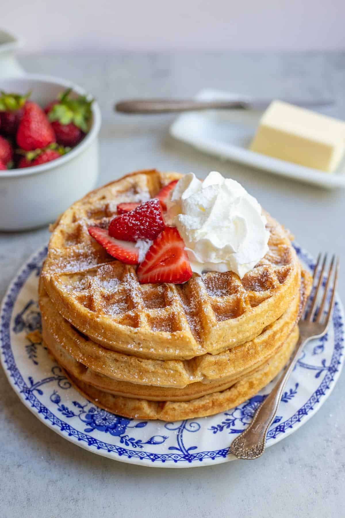 A stack of whole wheat waffles on a blue decorative plate topped with whipped cream and sliced strawberries.