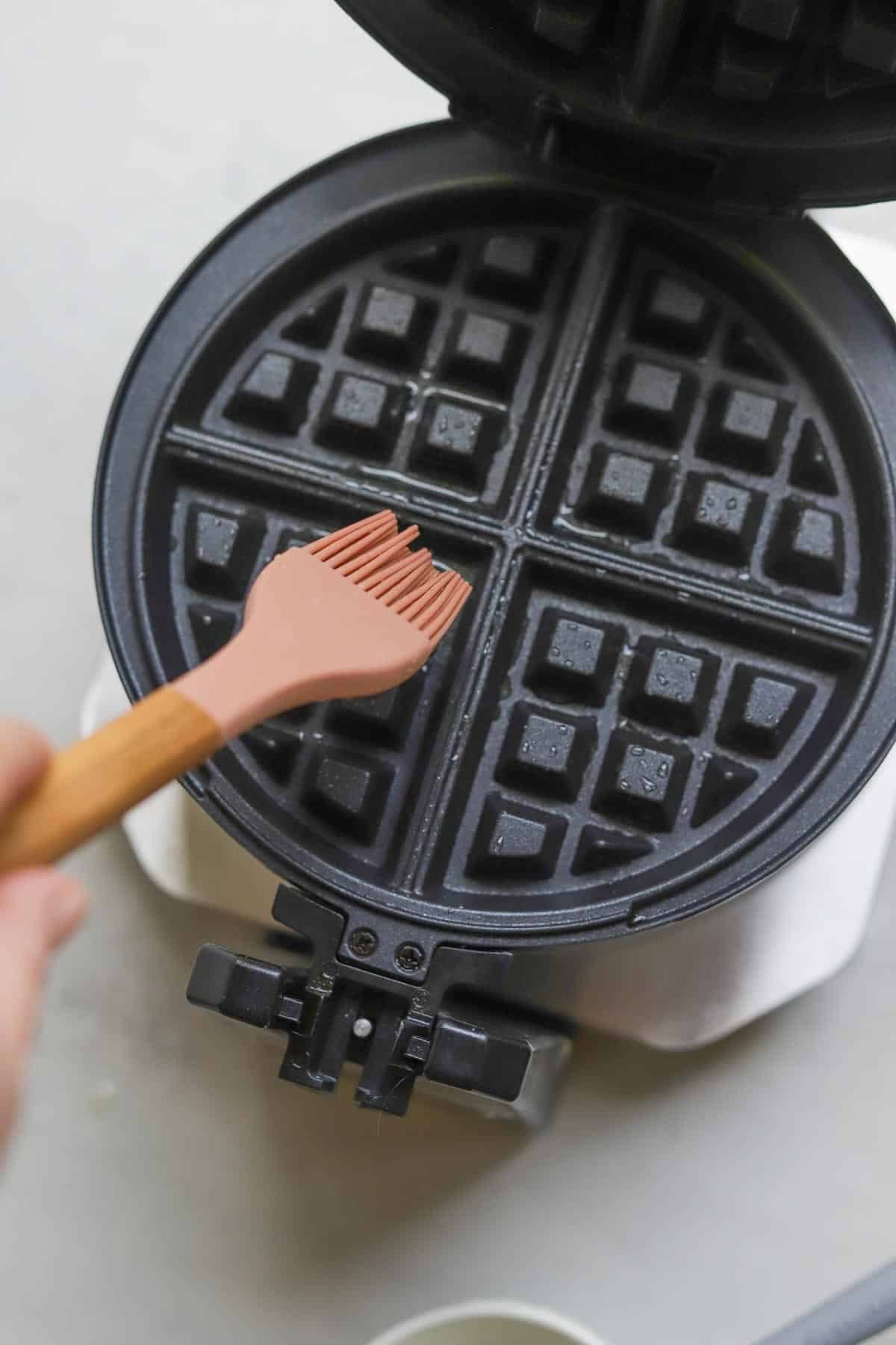 A silicone brush adding butter to a heated waffle maker.