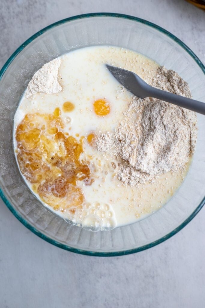 All the ingredients to make whole wheat waffles combined in a glass bowl with a spoon to stir the batter.