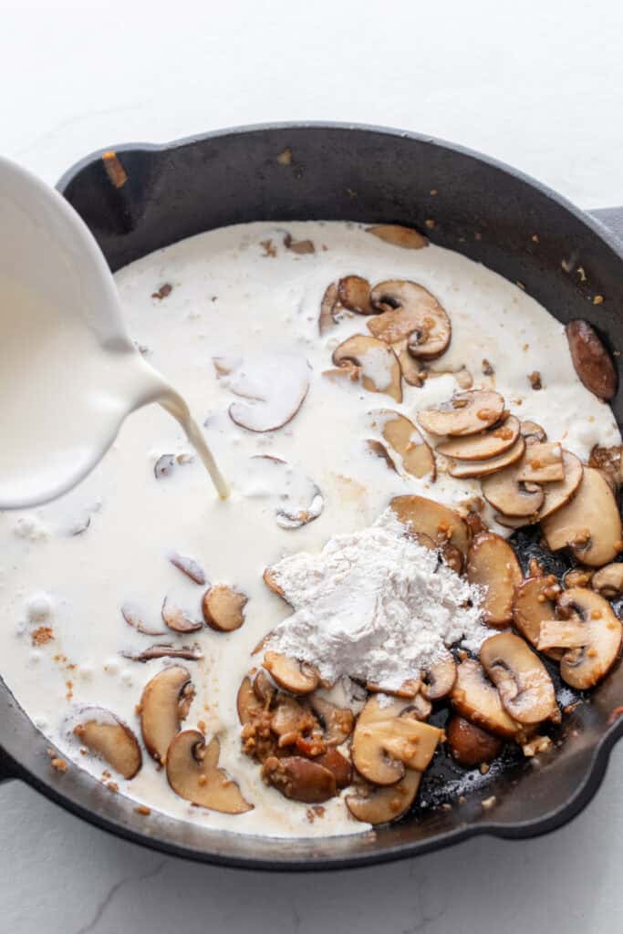 Pouring heavy cream into a cast iron skillet with browned mushrooms.