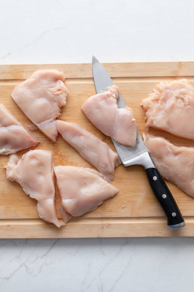 Six chicken breasts cut in half on a wooden cutting board with a knife.