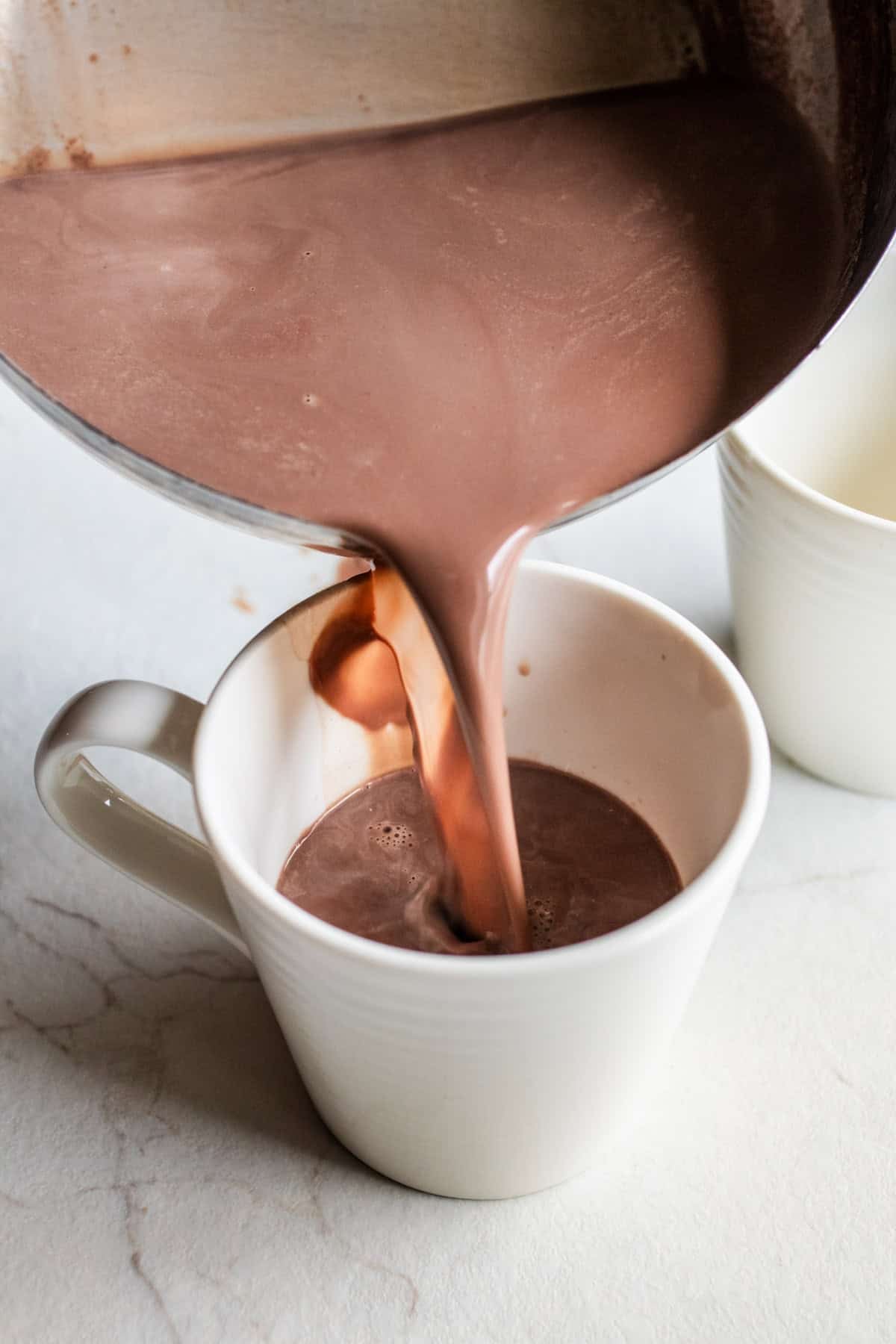 Hot chocolate being poured from a sauce pan into a white mug.