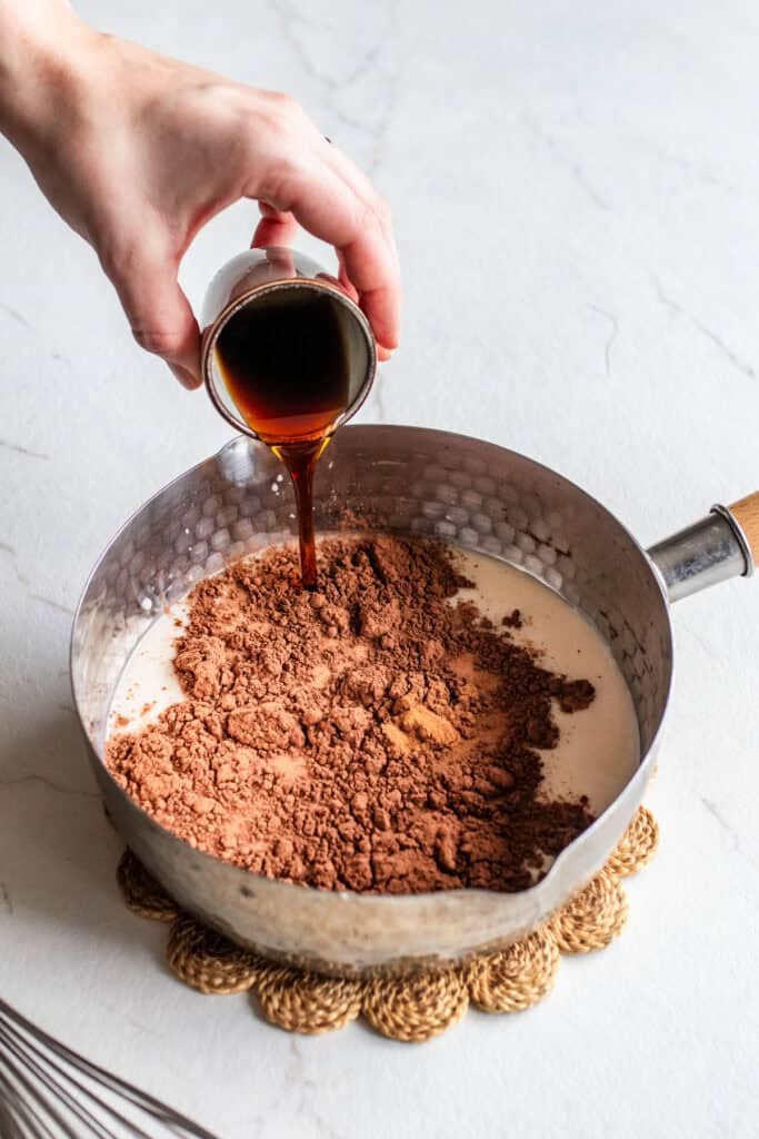 Maple syrup being added to a sauce pan with milk, cocoa powder, and bone broth.