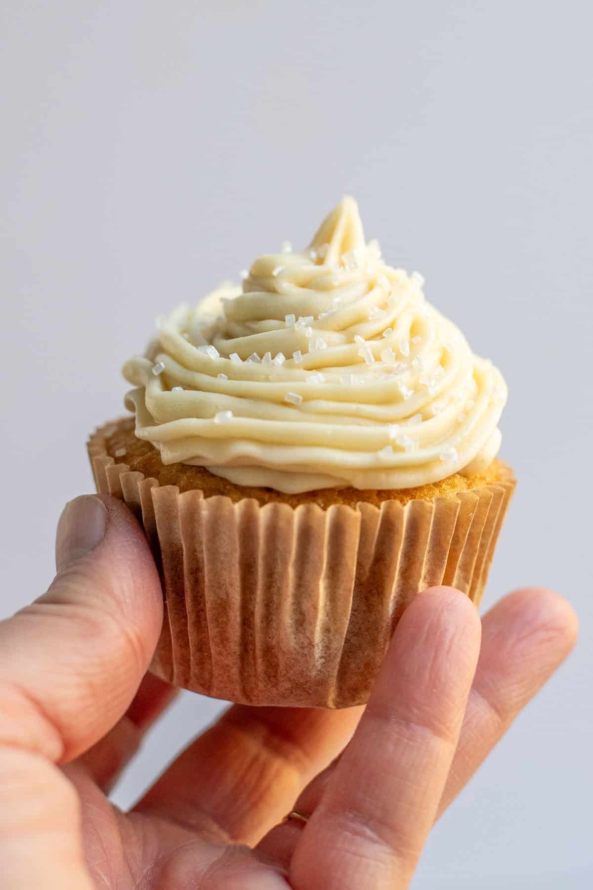 A woman's hand holding a vanilla cupcake piped with vanilla buttercream frosting.