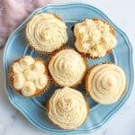 a blue plate with six vanilla cupcakes and piped with buttercream frosting in different designs.
