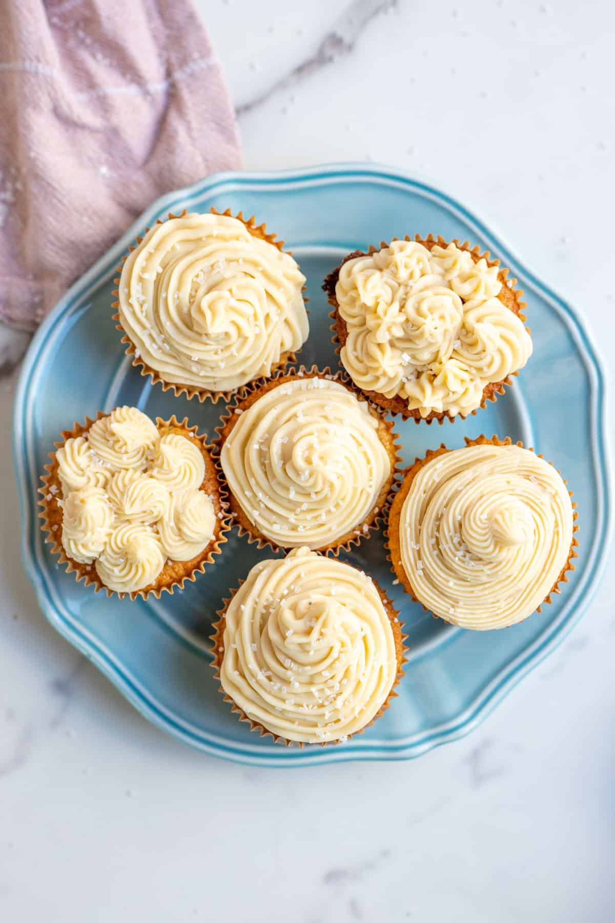 A blue plate with six vanilla cupcakes and piped with buttercream frosting in different designs.