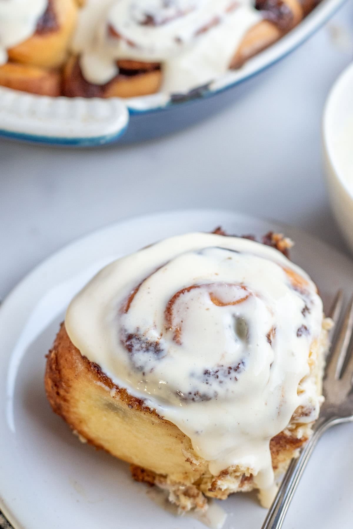One fluffy iced cinnamon roll on a white dessert plate with a fork to the side.