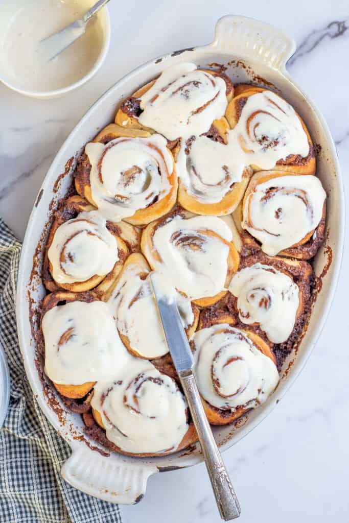 Freshly baked cinnamon rolls being frosted with a cream cheese glaze.