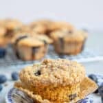 one whole wheat blueberry muffin on a blue decorative plate with a cooling wrack full of muffins in the background.