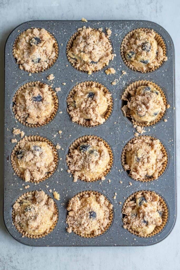 streusel topping sprinkled over blueberry muffin batter in the muffin tin.