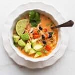 a bowl of southwest chicken soup topped with cheese, avocado, lime, and cilantro.