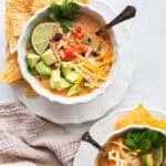 a bowl of chicken soup served with tortilla chips, lime, avocado, and cilantro.
