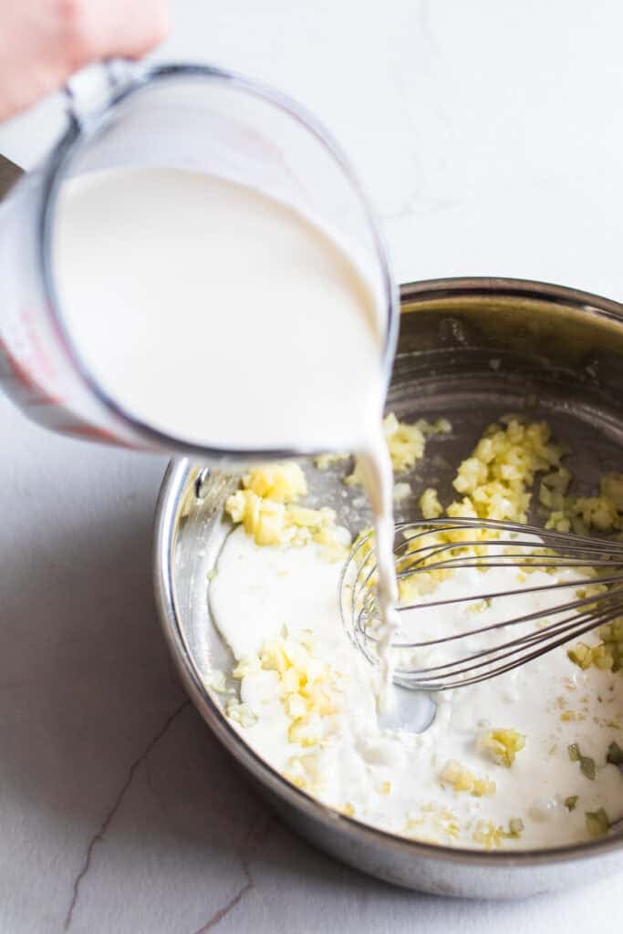 Cream being poured from a glass measuring cup into a sauce pan with onions and a whisk.