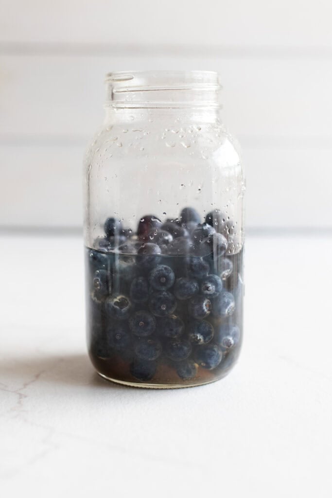 Blueberries added to liquid in a large mason jar on a counter top. 