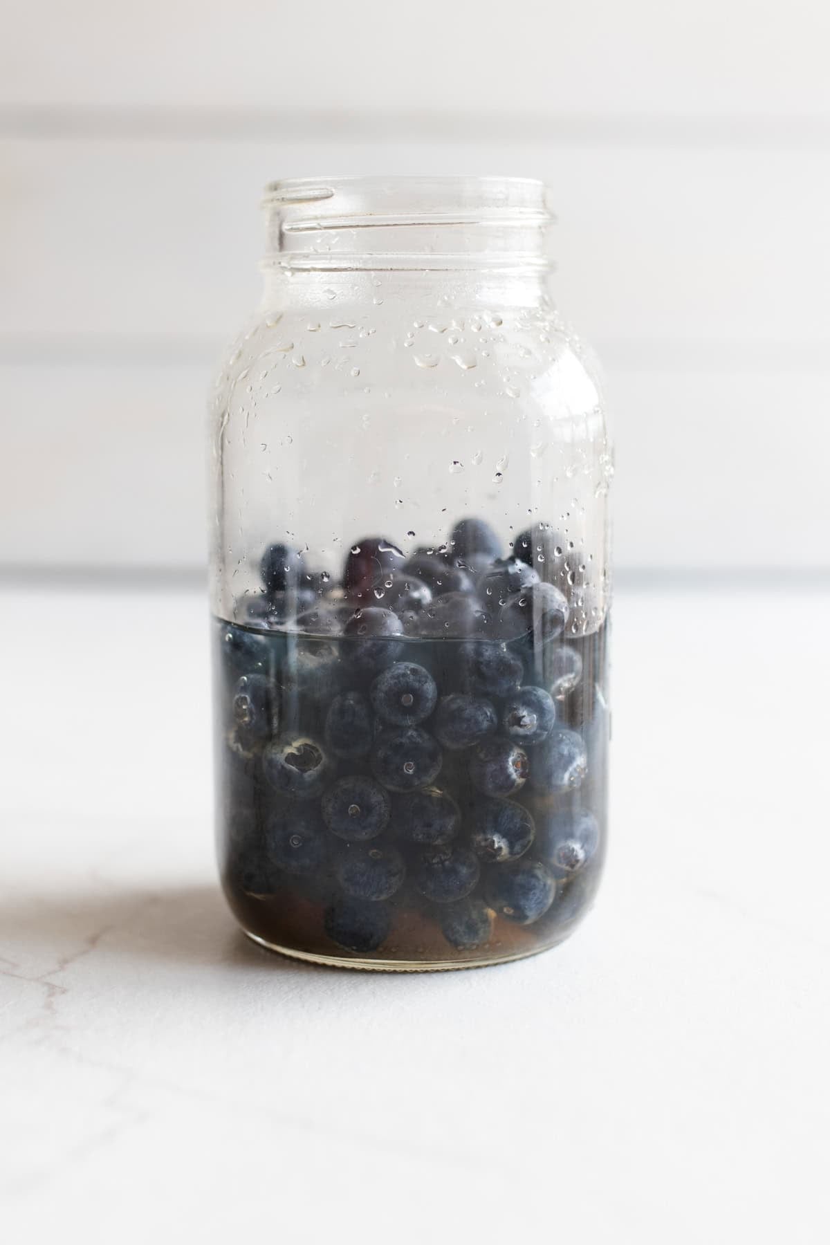 Blueberries added to liquid in a large mason jar on a counter top.