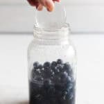 A hand adding a fermentation weight to a large mason jar full of liquid and blueberries.