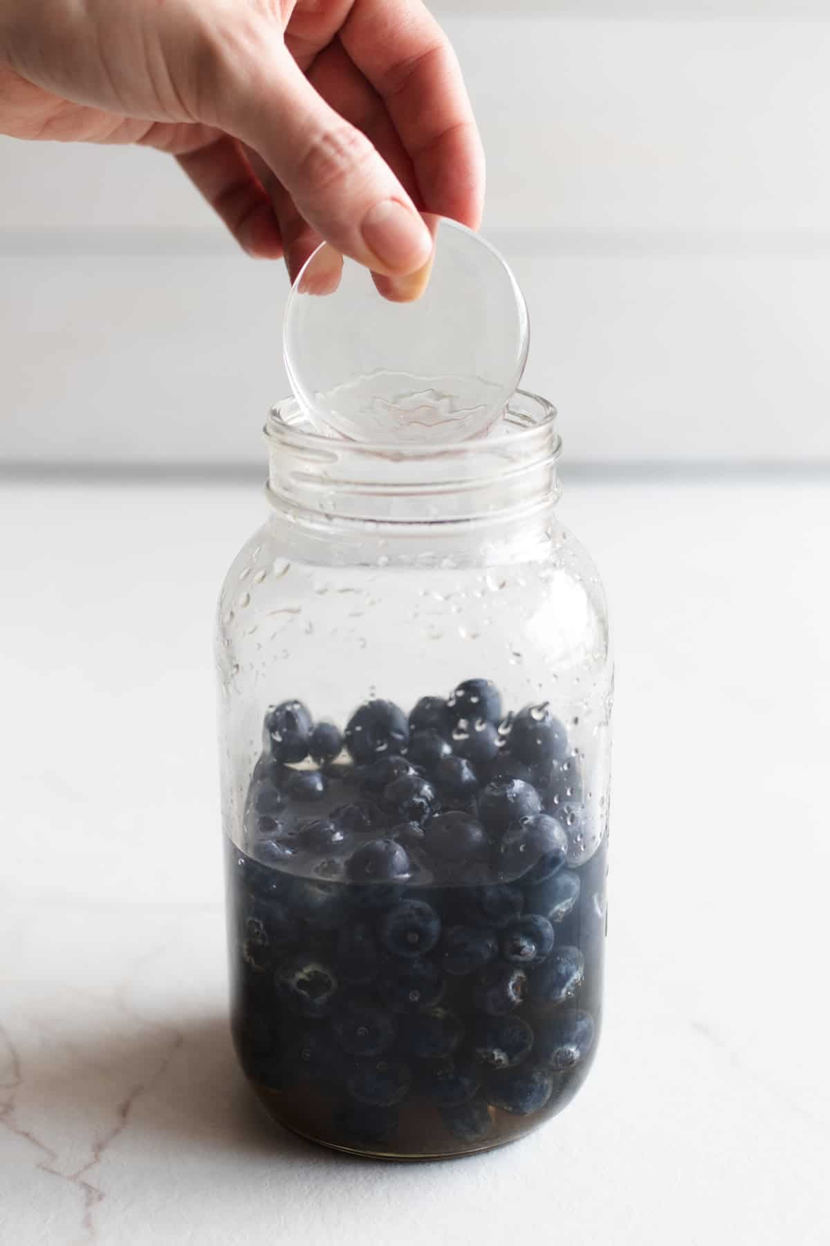 A hand adding a fermentation weight to a large mason jar full of liquid and blueberries.