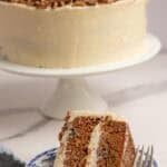 one slice of sourdough carrot cake on a blue decorative plate with the full cake on a white cake plate in the background.