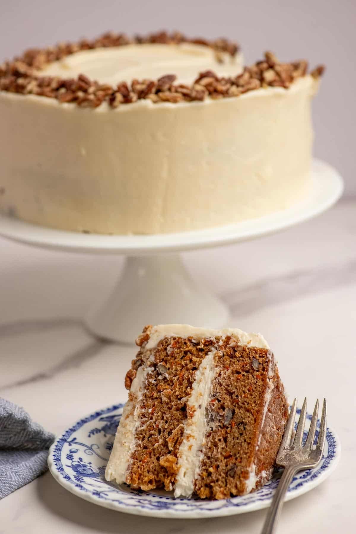one slice of sourdough carrot cake on a blue decorative plate with the full cake on a white cake plate in the background.