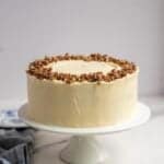 a frosted carrot cake on a white cake plate topped with chopped pecans.