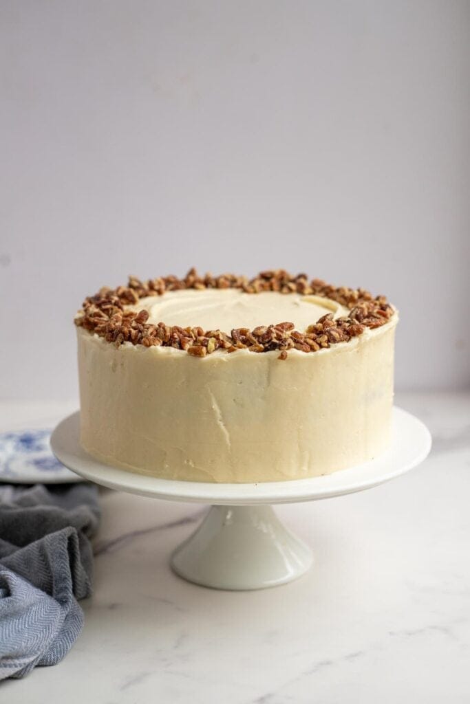 a frosted carrot cake on a white cake plate topped with chopped pecans. 