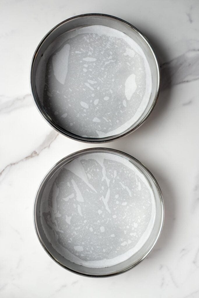 two round cake pans that are lined with parchment paper on a marble countertop.