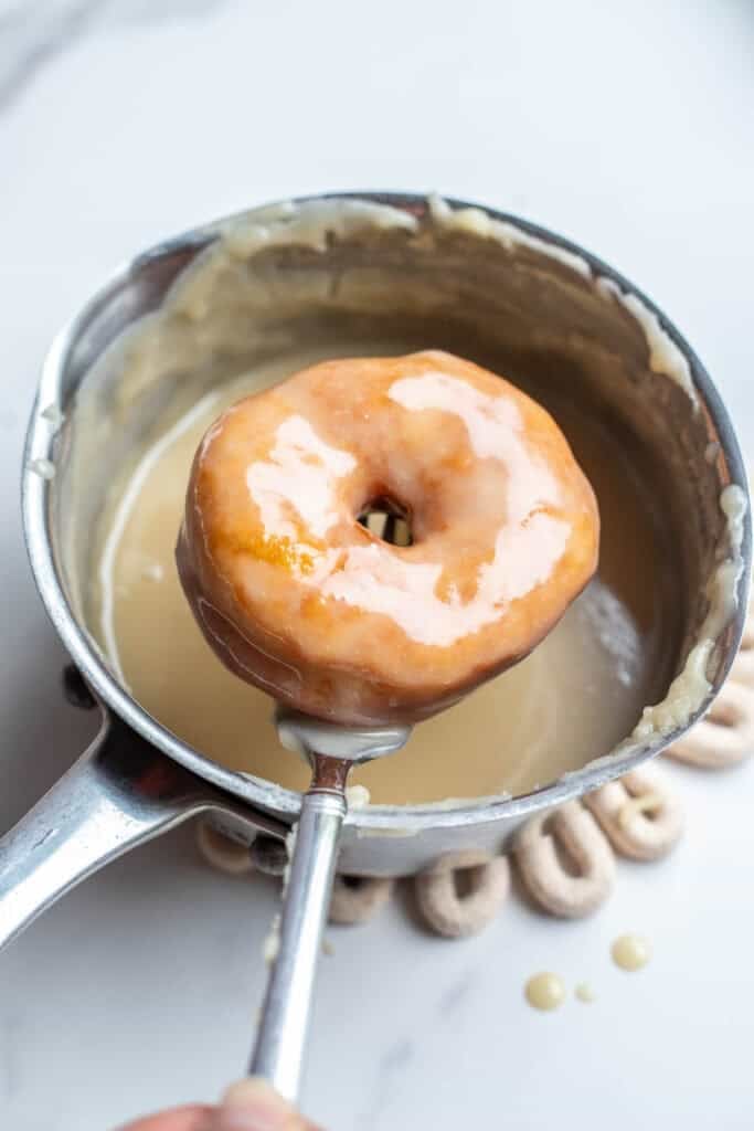 A sourdough donut dipped into a pot of vanilla glaze. 
