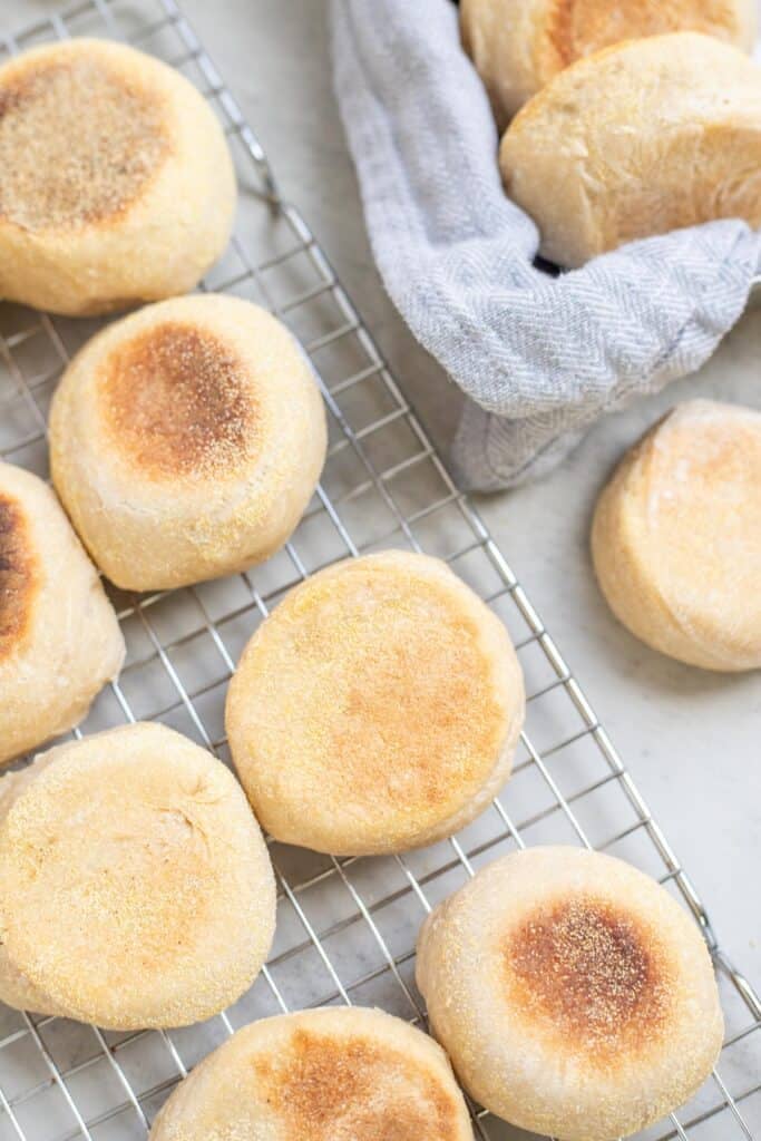 Browned English muffins cooling on a wire rack on the kitchen counter.