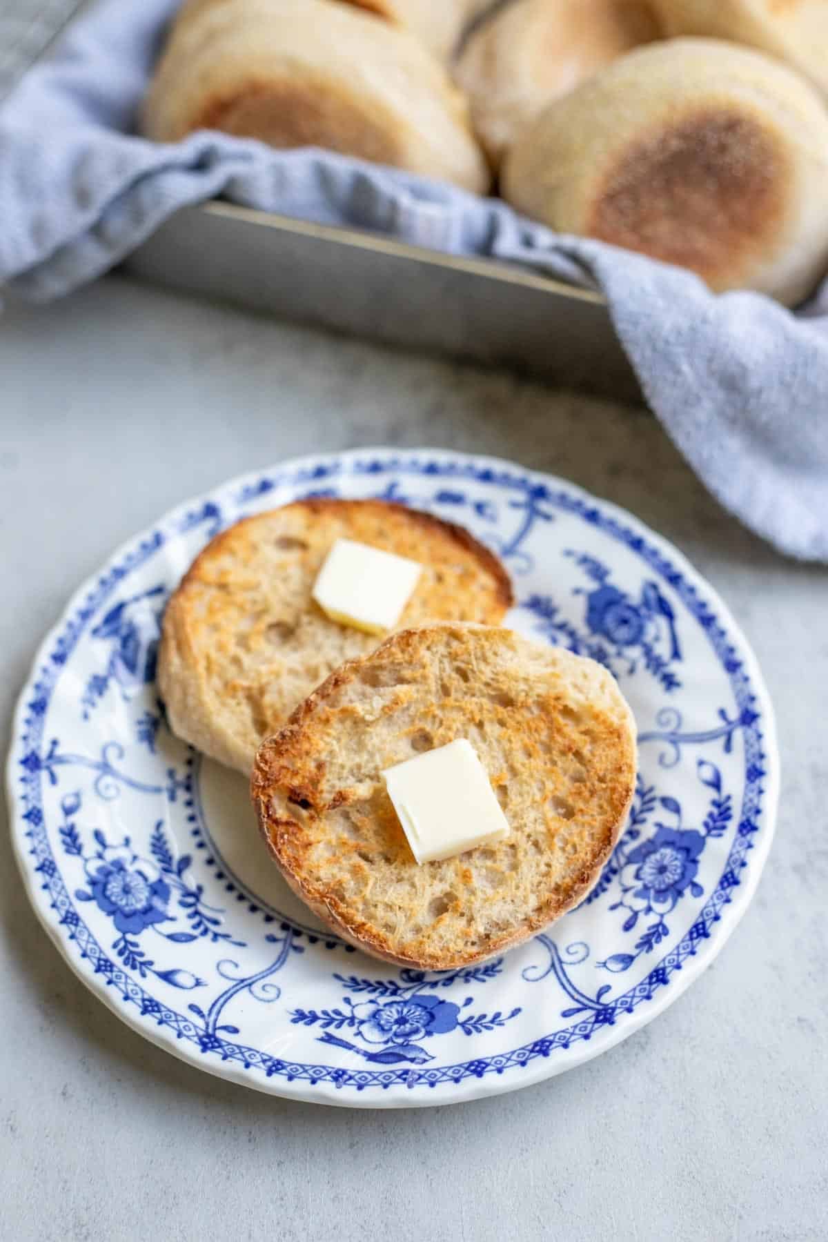 An english muffin split open with a pad of butter on each side of the muffin on a blue decorative plate.