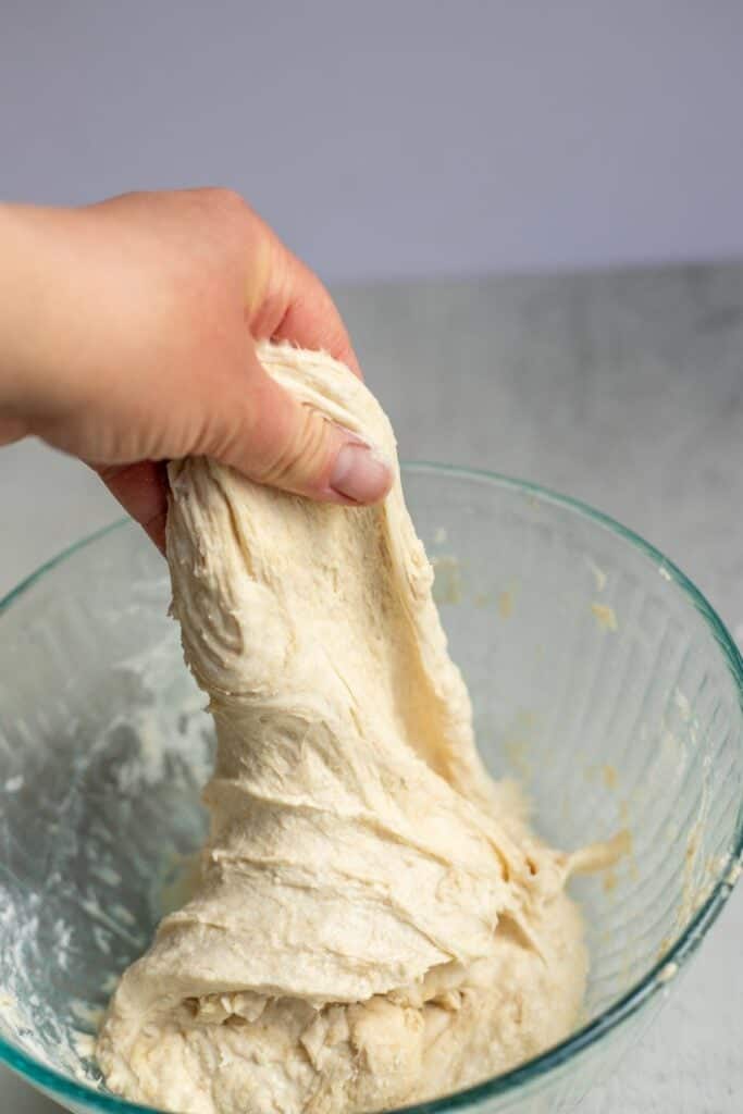 A hand reaching in a glass bowl and stretching sourdough upward in a series of stretch and folds.