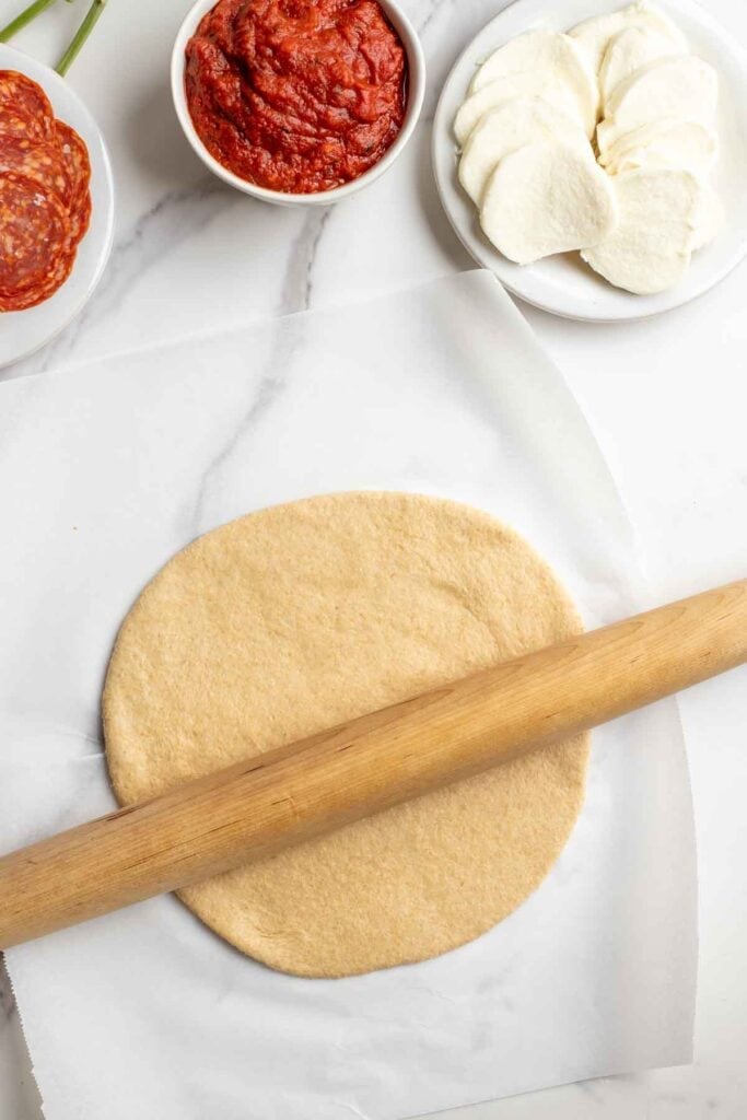Pizza dough rolled out on a white countertop with a rolling pin on top of the dough and bowls of pizza toppings on the side.