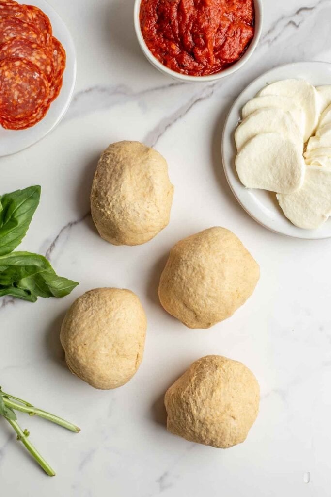 Four small dough balls on a countertop next to a plate of pizza toppings.
