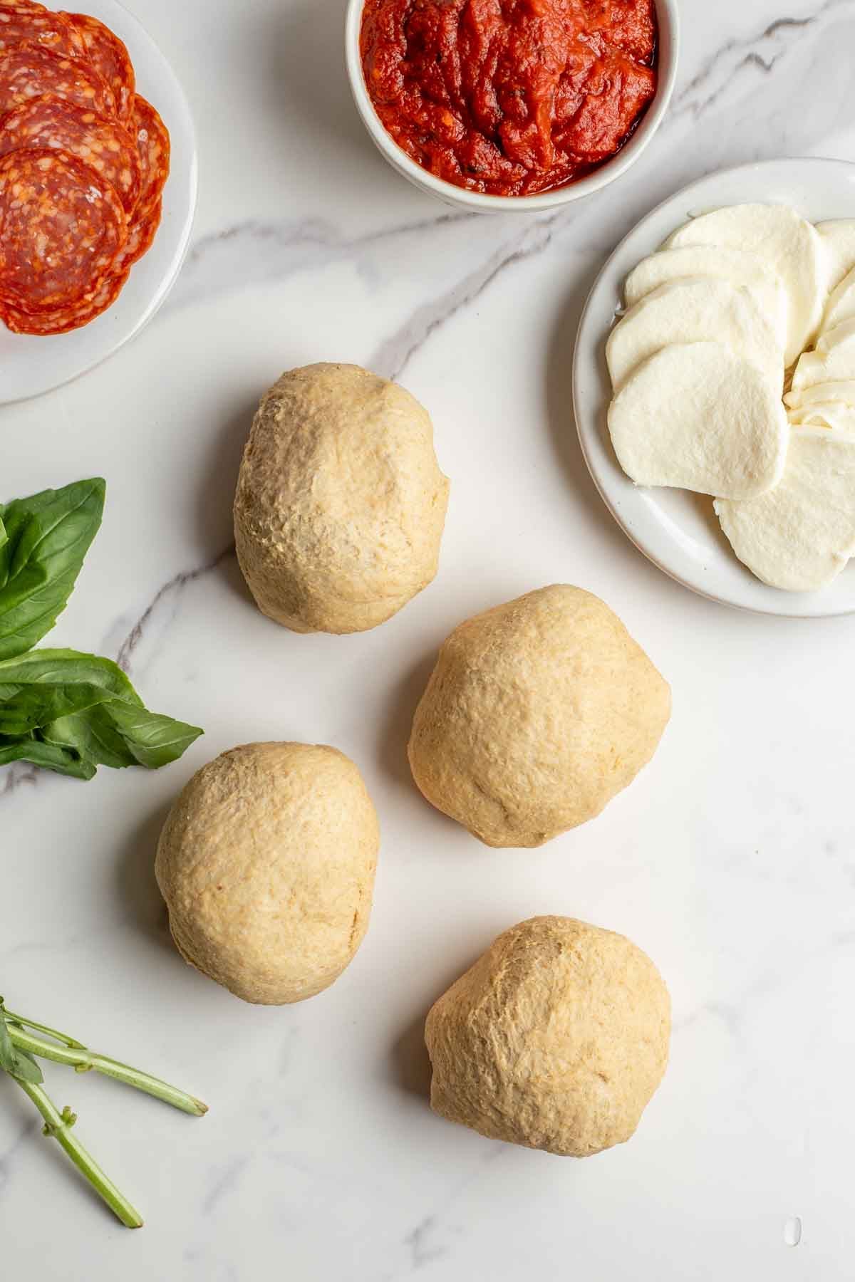 Four small dough balls on a countertop next to a plate of pizza toppings.