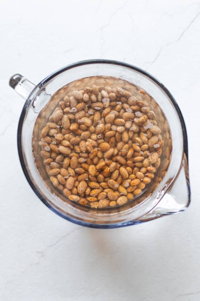 Pinto beans soaking in water in a large glass measuring cup.