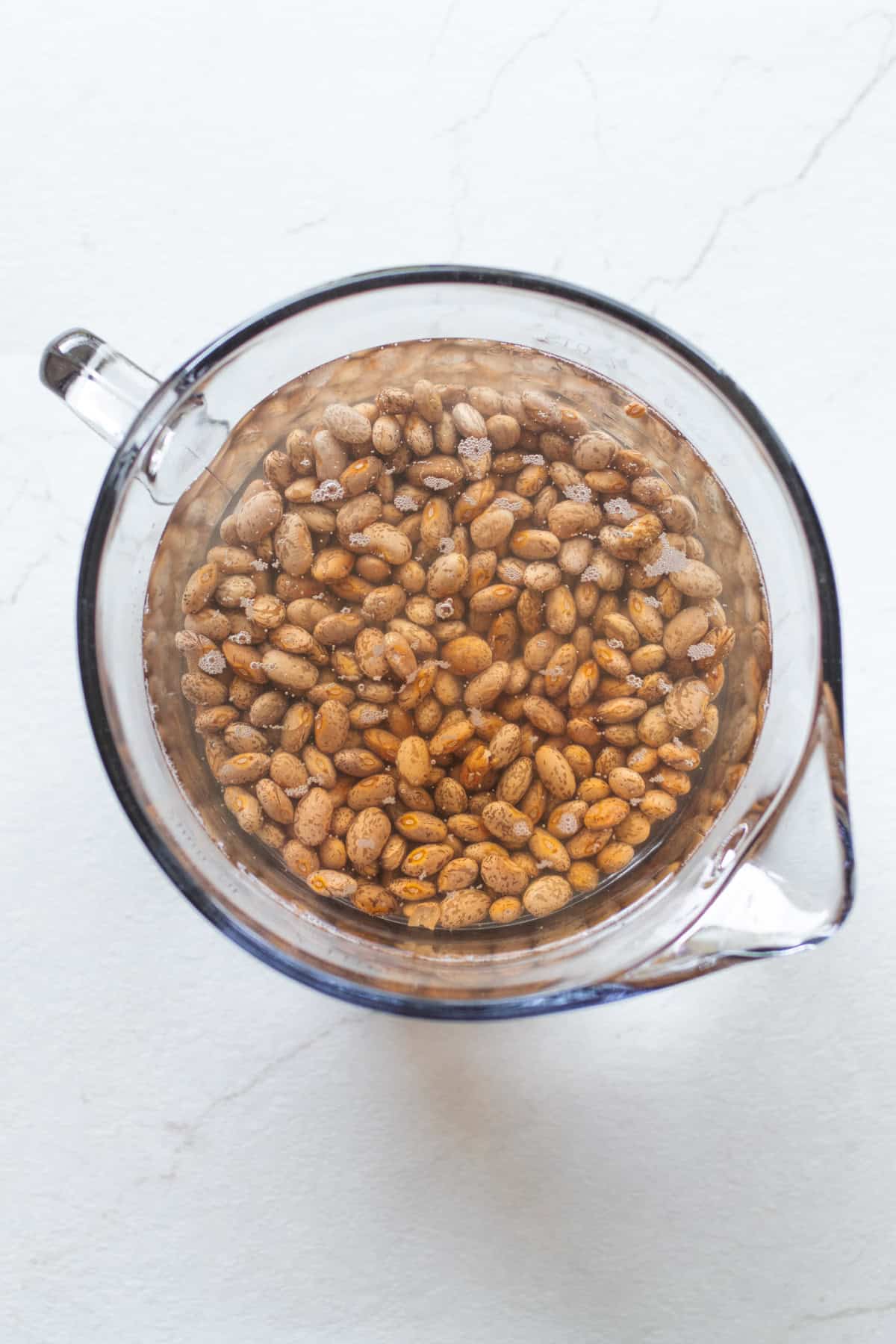 Pinto beans soaking in water in a large glass measuring cup.