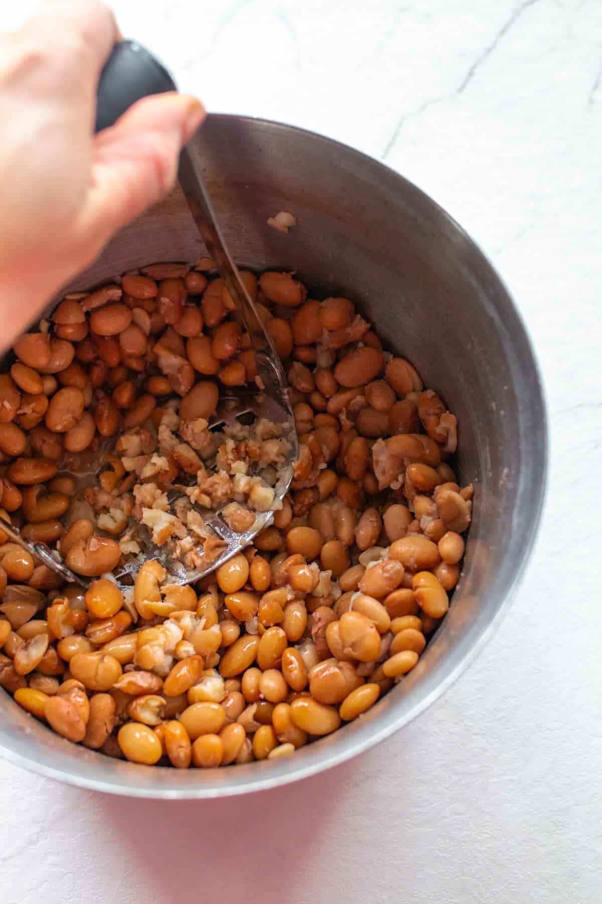 Cooked pinto beans in a sauce pan with a hand using a potato masher in them.